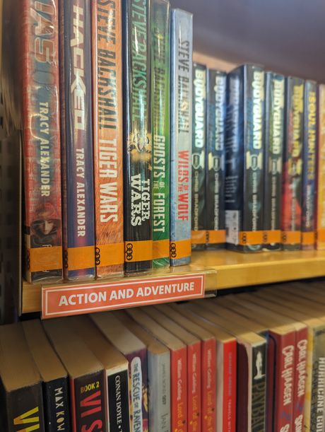 A bookshelf with a card sign sat flush to the shelf. It is on an orange background, with a white border and white text on it reading 'Action and Adventure'. It is sleek and integrated with the bookshelf.